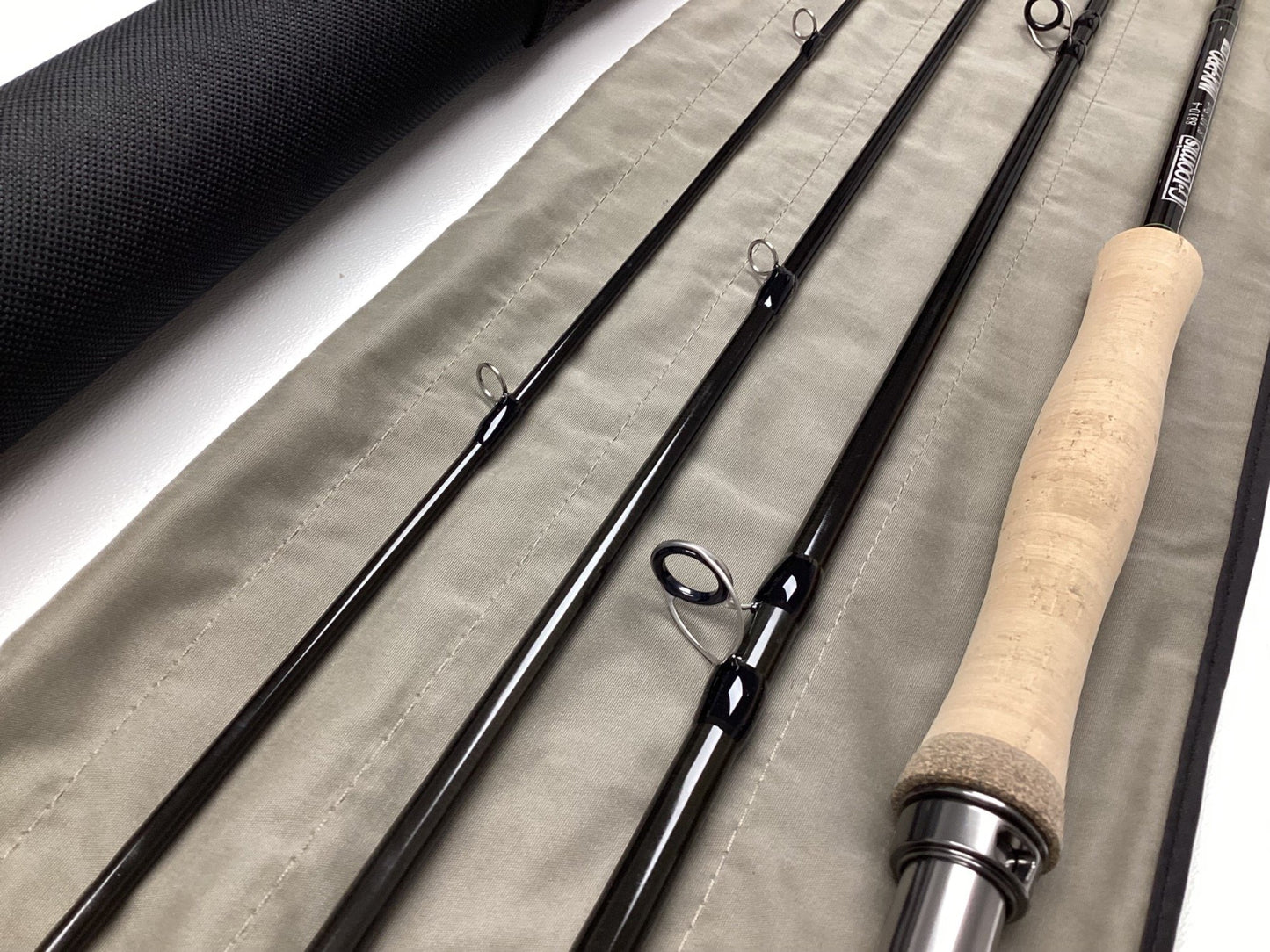 A close-up of the G. Loomis IMX-PRO V2 Fly Rod 8810-4, a used fly rod in mint condition, shows its cork handle and metal guides on a tan case beside a black tube—ideal for upgrading with our rod and reel trade-in program.