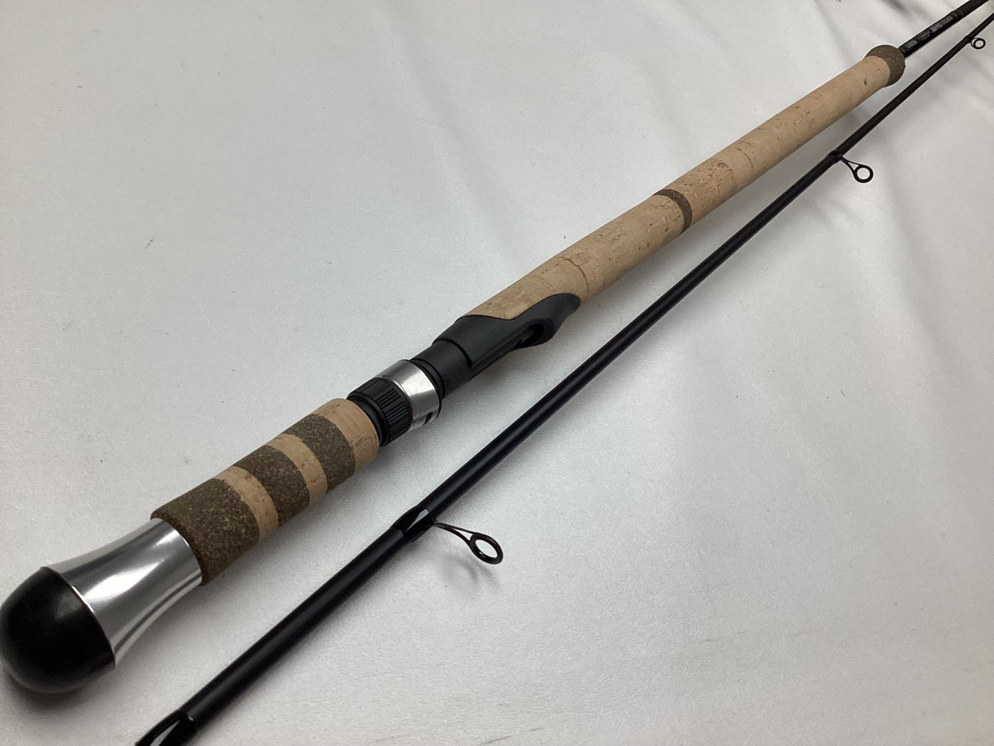 A close-up of a G. Loomis IMX-PRO Steelhead Centerpin Rod 1361-2CP STR 11'4" Medium Light (Used-Mint) shows its cork handle, metal reel seat, and black segments on a white surface.