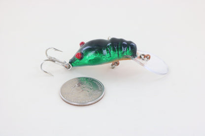 The WE Baits Red Eye Craw Green Foil fishing lure by WE Baits, featuring green foil, black accents, red eyes, and a clear plastic lip, is shown next to a U.S. quarter for size comparison on a white background.