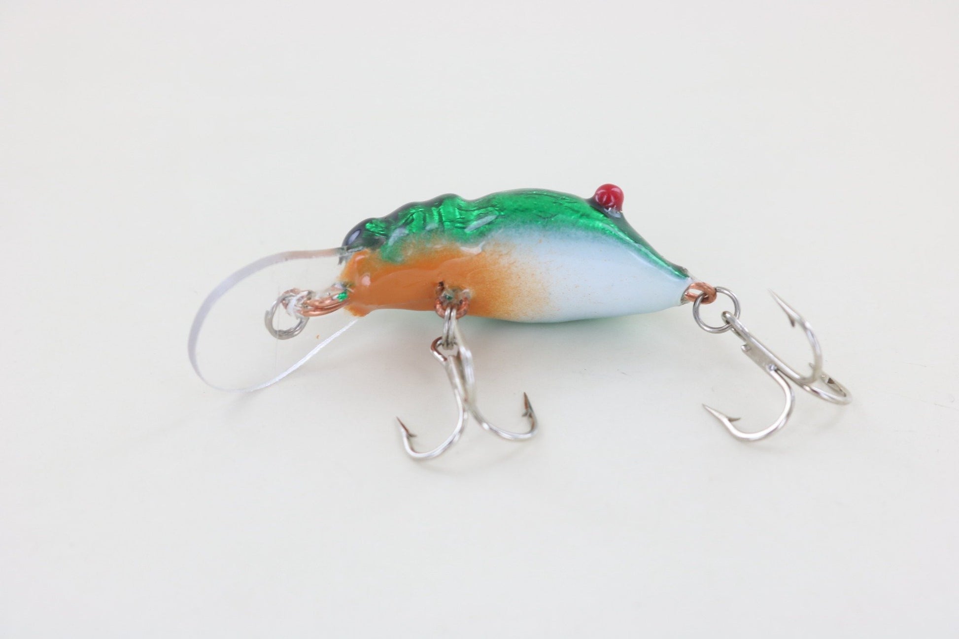 The WE Baits Red Eye Craw Green Foil by WE Baits is a fishing lure with a green top, orange middle, and white underside. It features two treble hooks and a clear plastic lip and is shown on a plain white background.