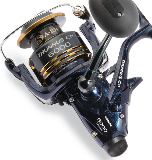 A close-up of the Shimano Thunnus 6000CI4 Saltwater Spinning Reel by Shimano, showcasing its black and gold spool, ergonomic handle, detailed branding, and sleek metallic blue frame.