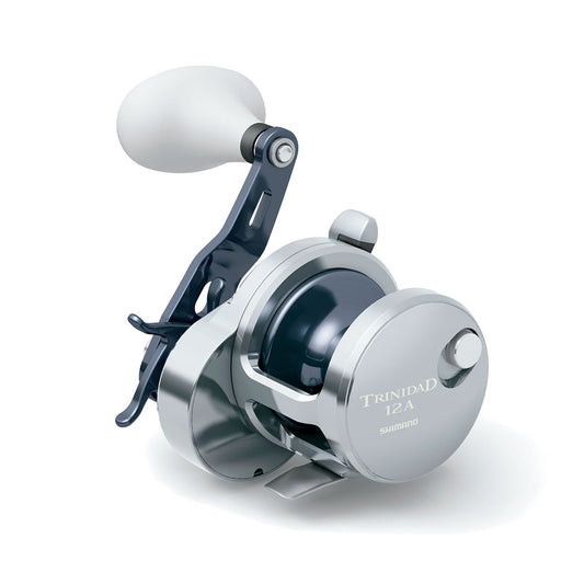 The Shimano Trinidad 12A Conventional Reel by Shimano features High Efficiency Gearing, a large white handle, and a silver finish with an etched logo. Shown on a white background, it’s perfect for serious saltwater anglers.